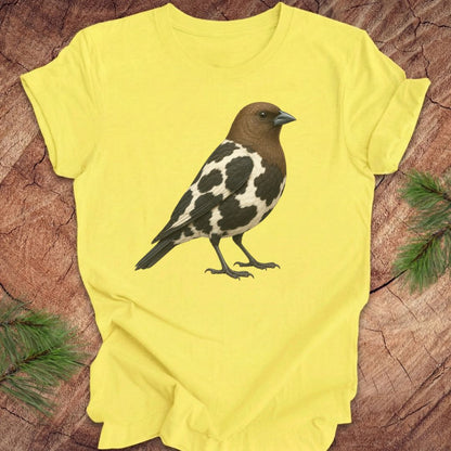 A bright yellow short-sleeve t-shirt laid flat on a textured wood background, featuring a detailed graphic of a brown, black, and white speckled bird on the front.