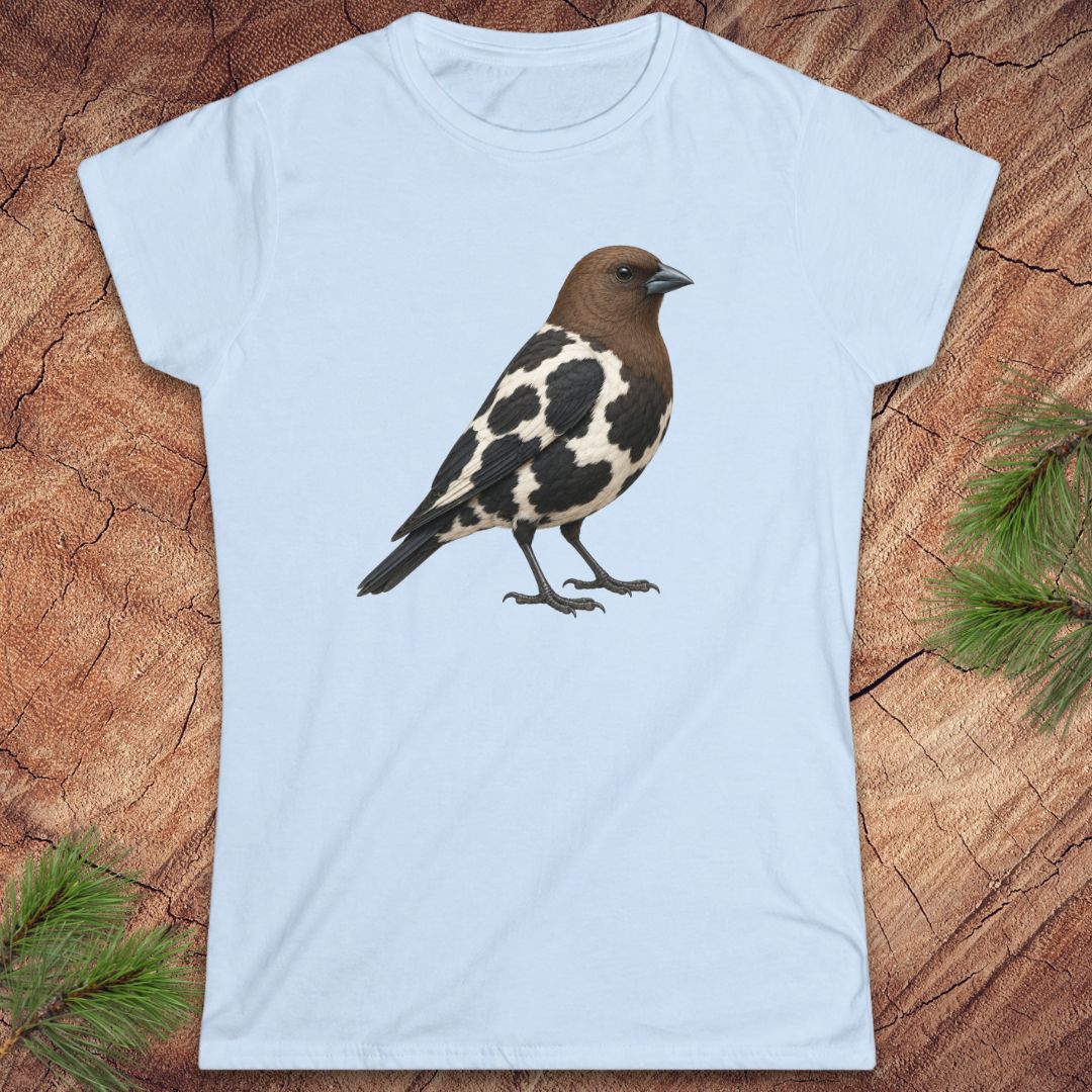 A light blue short-sleeve crewneck t-shirt featuring a graphic print of a brown and black-and-white speckled bird, displayed flat on a textured wood background with pine needles.