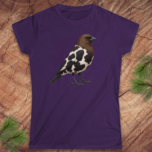A purple short-sleeve crewneck t-shirt featuring a graphic of a black and white speckled bird with a brown head, laid flat on a textured wood background with pine needles in the corners.