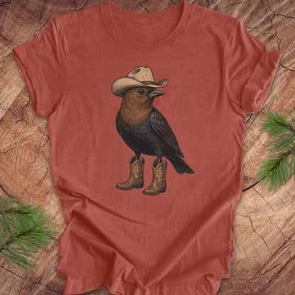 A terracotta-colored short-sleeve t-shirt featuring a graphic of a brown and black bird wearing a beige cowboy hat and matching cowboy boots, laid flat on a rustic wood background with pine needles in the corners.