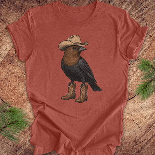 A terracotta-colored short-sleeve t-shirt featuring a graphic of a brown and black bird wearing a beige cowboy hat and matching cowboy boots, laid flat on a rustic wood background with pine needles in the corners.