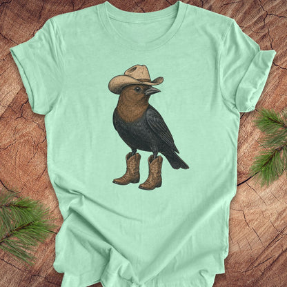 A mint green short-sleeve t-shirt laid flat on a rustic wood surface, featuring a graphic of a black bird wearing a tan cowboy hat and brown cowboy boots.
