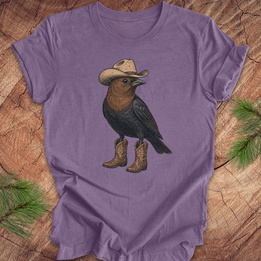 A purple short-sleeve t-shirt laid flat on a rustic wood surface, featuring a graphic of a black bird wearing a beige cowboy hat and brown boots.