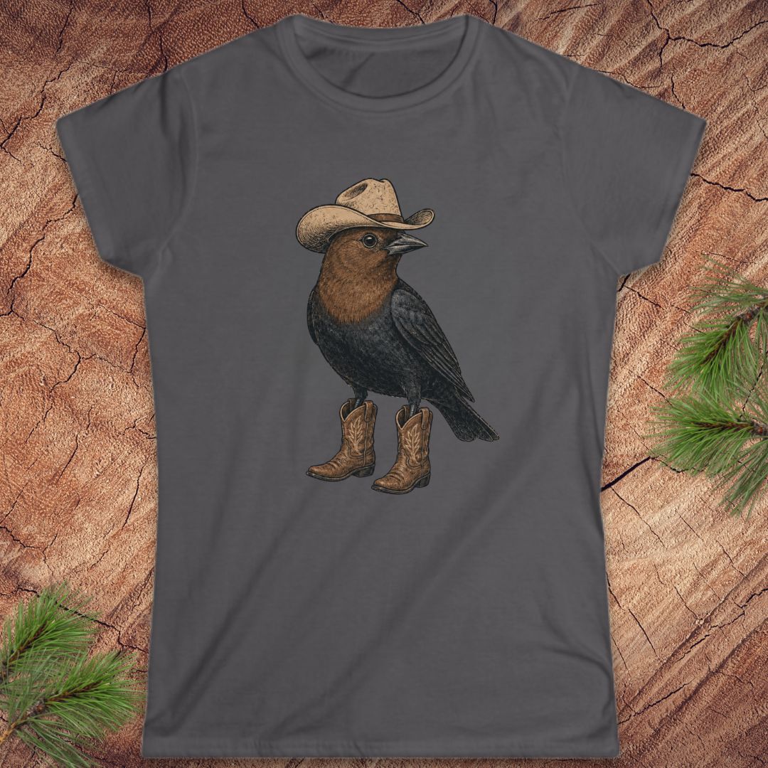 A dark gray women's short-sleeve t-shirt featuring a graphic of a black bird wearing a beige cowboy hat and brown cowboy boots, laid flat on a rustic wood background with pine needles.