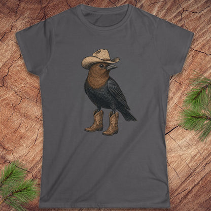 A dark gray women's short-sleeve t-shirt featuring a graphic of a black bird wearing a beige cowboy hat and brown cowboy boots, laid flat on a rustic wood background with pine needles.