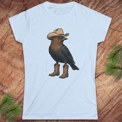A light blue short-sleeve crewneck t-shirt featuring a graphic of a brown bird wearing a cowboy hat and boots, laid flat on a textured wood background with pine needles in the corners.
