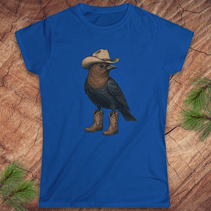 A royal blue short-sleeve t-shirt laid flat on a textured wood background with pine branches in the corners. The shirt features a graphic of a black bird wearing a beige cowboy hat and brown cowboy boots.