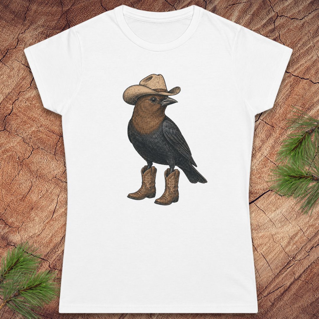A white short-sleeve crewneck t-shirt featuring a graphic of a brown and black bird wearing a tan cowboy hat and brown cowboy boots, displayed flat on a textured wood background with pine needles.