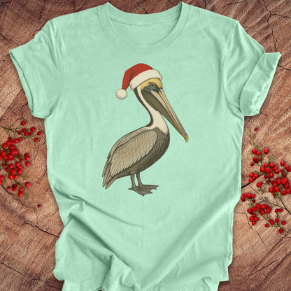 A mint green short-sleeve t-shirt featuring a graphic of a brown pelican wearing a red Santa hat, laid flat on a rustic wood background decorated with red berries.