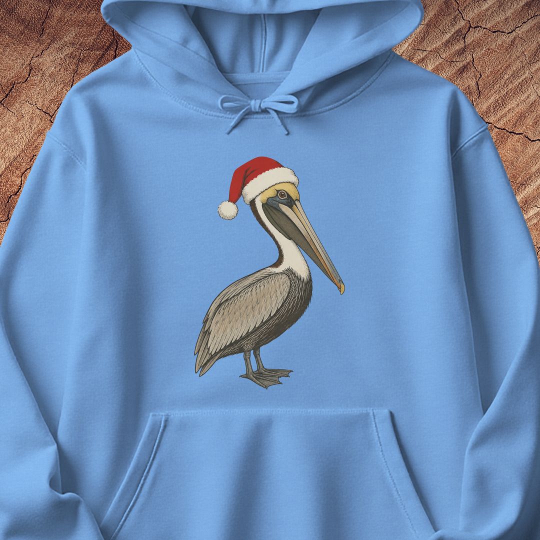 A light blue hoodie laid flat on a rustic wood background, featuring a printed illustration of a brown pelican wearing a red Santa hat on the chest.
