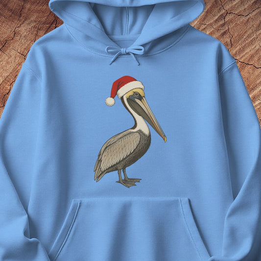 A light blue hoodie laid flat on a rustic wood background, featuring a printed illustration of a brown pelican wearing a red Santa hat on the chest.