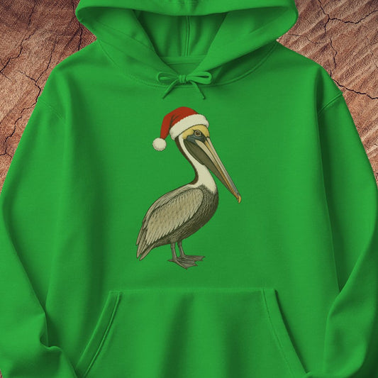 A bright green pullover hoodie laid flat on a textured brown wood background, featuring a printed illustration of a brown pelican wearing a red Santa hat on the chest.