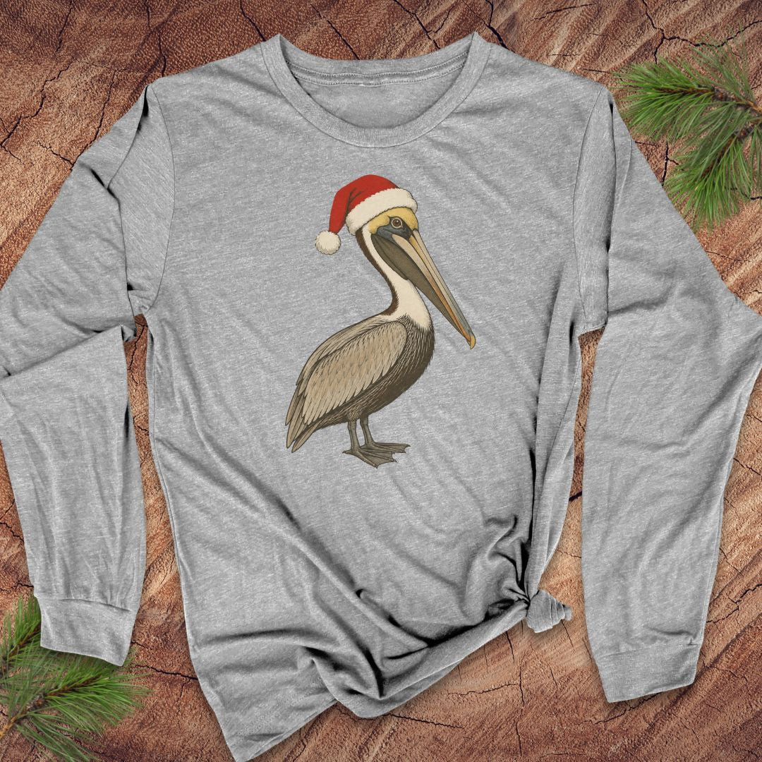 A heather gray long-sleeve t-shirt featuring a graphic of a brown pelican wearing a red Santa hat, laid flat on a rustic wood background with pine needles.