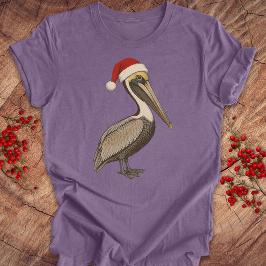 A purple short-sleeve t-shirt laid flat on a rustic wood background, featuring a graphic of a brown pelican wearing a red Santa hat. Red berry sprigs are placed on both sides of the shirt.