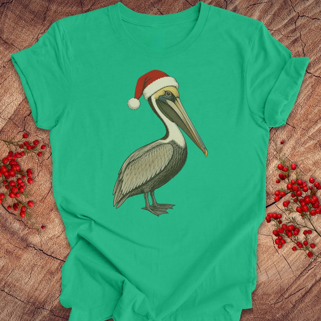 A green short-sleeve crewneck t-shirt featuring a graphic of a brown pelican wearing a red Santa hat, laid flat on a rustic wood background with red berries.