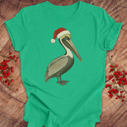 A green short-sleeve crewneck t-shirt featuring a graphic of a brown pelican wearing a red Santa hat, laid flat on a rustic wood background with red berries.