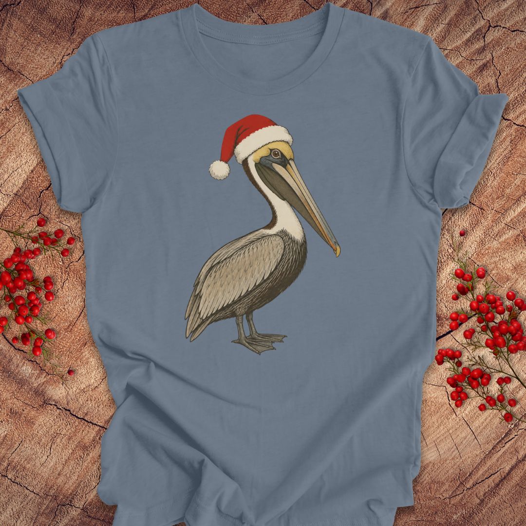 A dusty blue crewneck t-shirt featuring a graphic of a brown and white pelican wearing a red Santa hat, displayed on a rustic wood background with red berries.