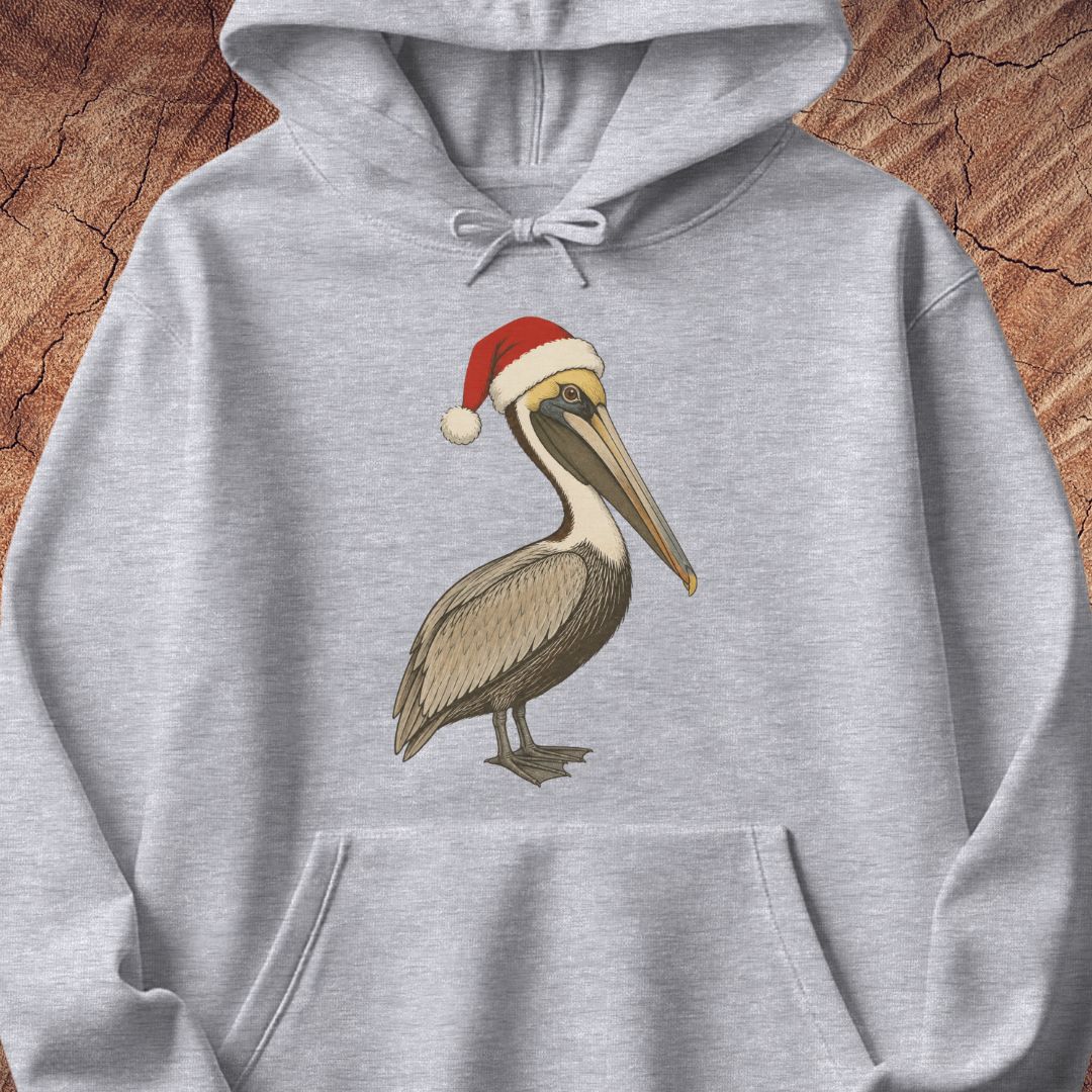 A light heather gray pullover hoodie featuring a printed illustration of a brown pelican wearing a red Santa hat with a white pom-pom, displayed against a rustic wood background.
