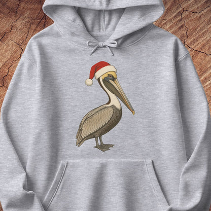 A light heather gray pullover hoodie featuring a printed illustration of a brown pelican wearing a red Santa hat with a white pom-pom, displayed against a rustic wood background.
