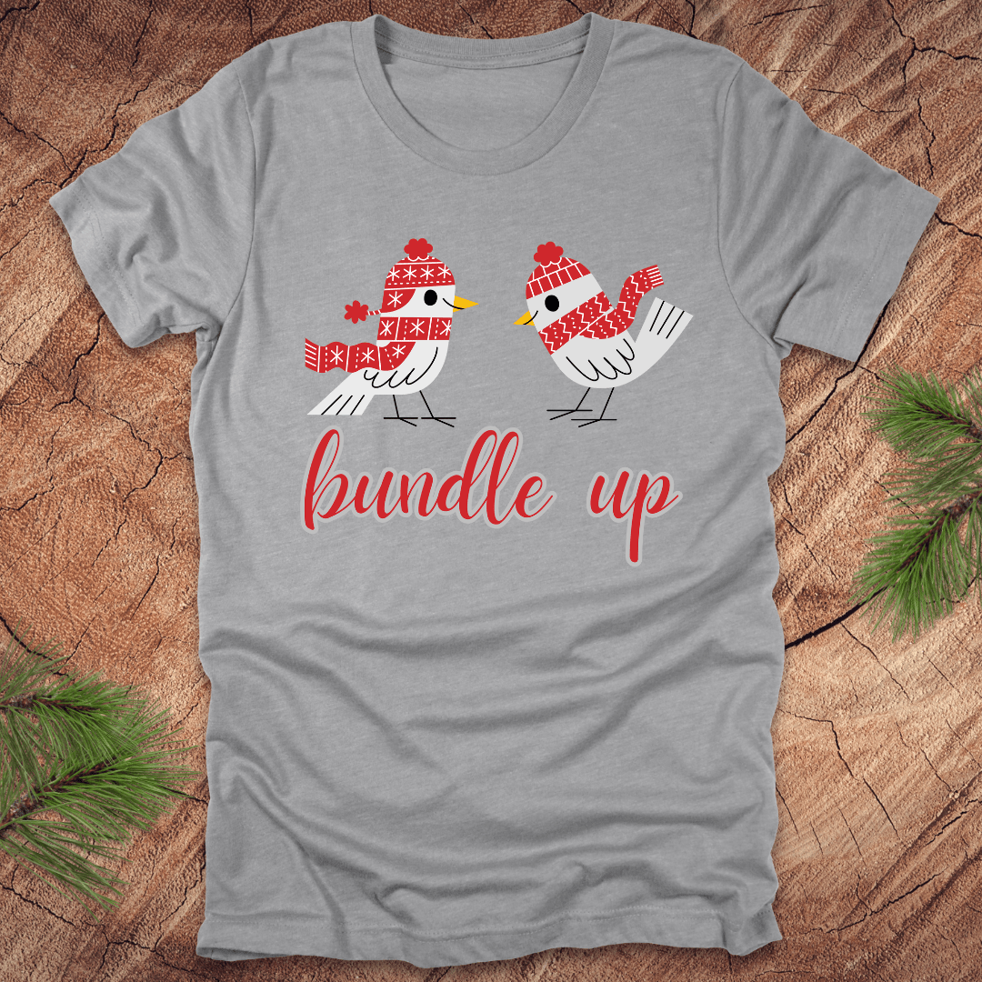Grey t-shirt with two birds with hats and scarves and text, 'Bundle up' on a wood background