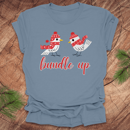 Light blue t-shirt with two birds with hats and scarves and text, 'Bundle up' on a wood background