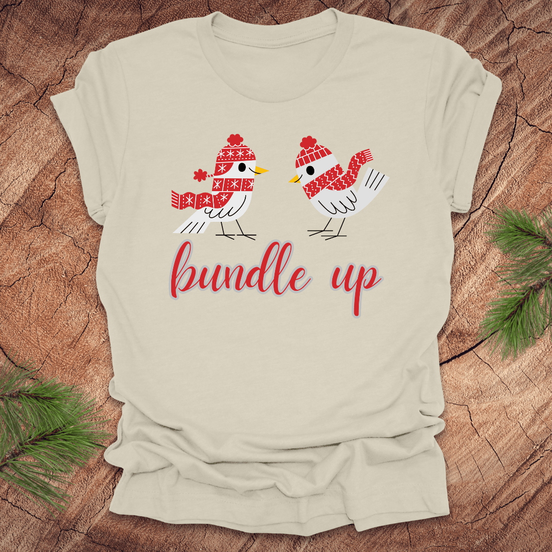Beige t-shirt with two birds with hats and scarves and text, 'Bundle up' on a wood background