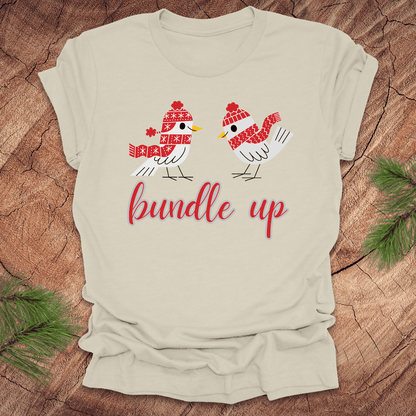 Beige t-shirt with two birds with hats and scarves and text, 'Bundle up' on a wood background