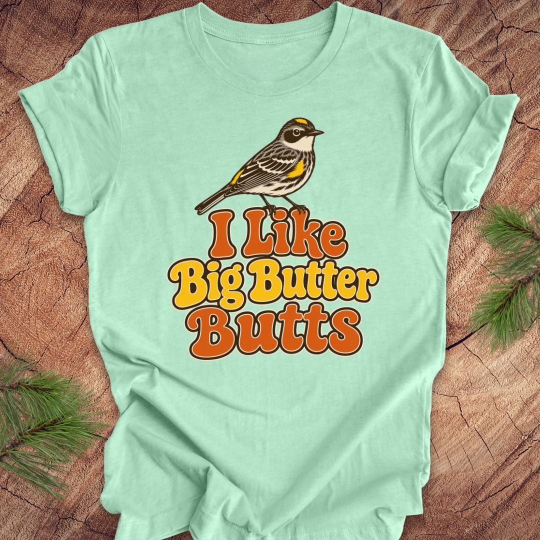 Light green t-shirt with a bird graphic and text 'I Like Big Butter Butts' on a wooden surface.