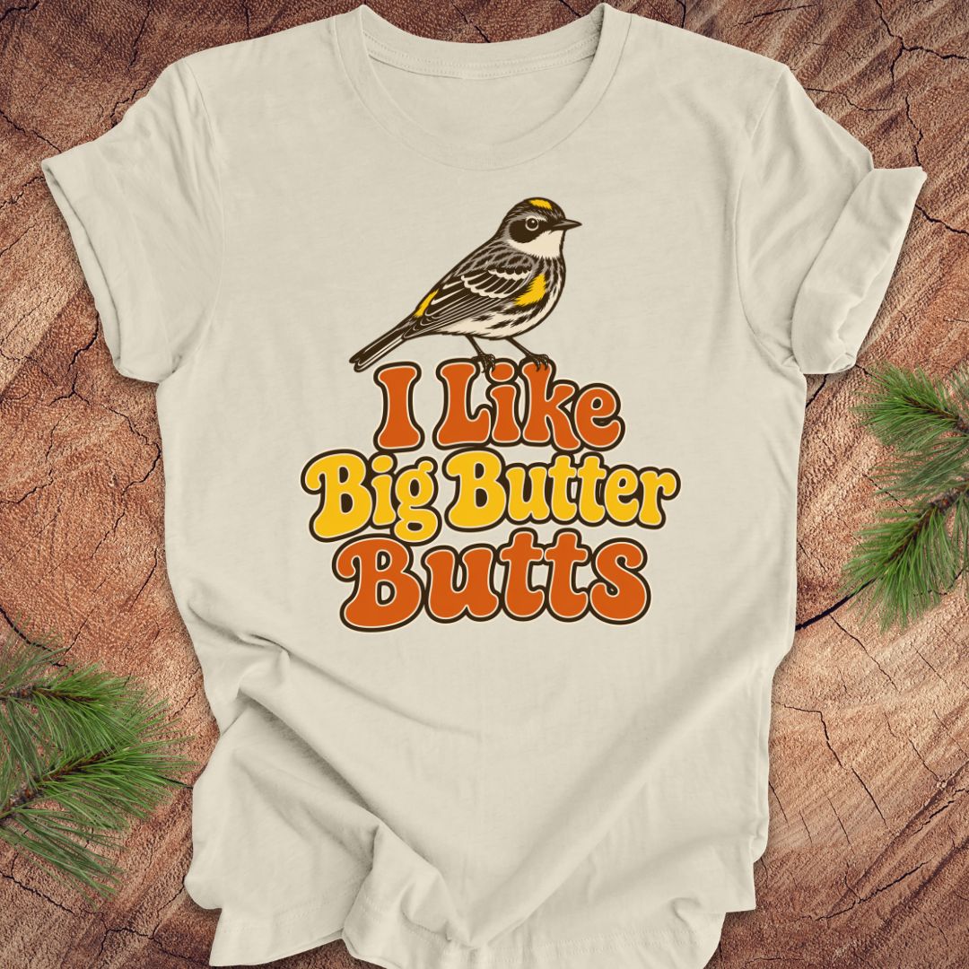 Beige t-shirt with a bird graphic and text 'I Like Big Butter Butts' on a wooden surface.