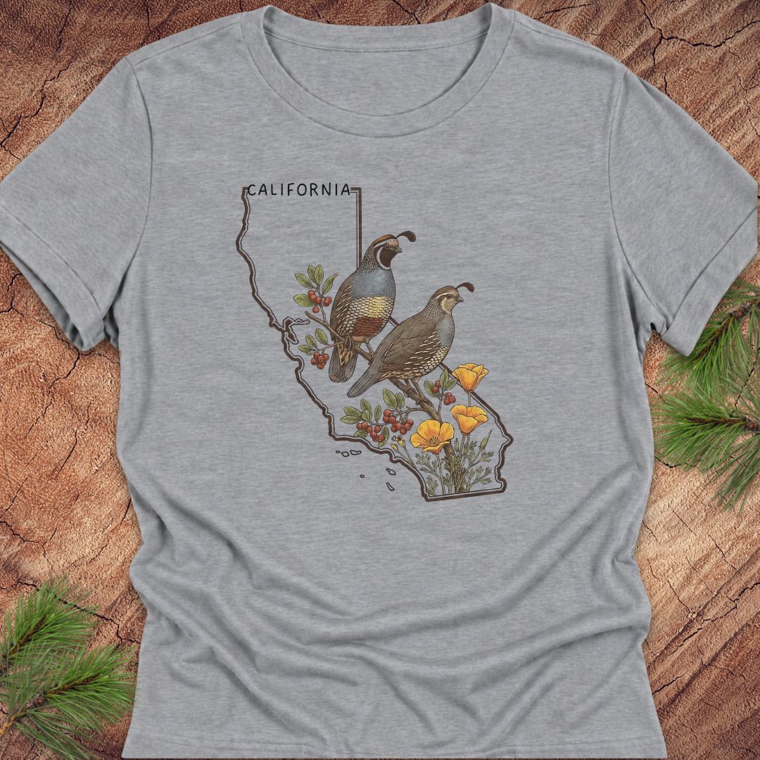 A heather gray short-sleeve t-shirt with a graphic print of two quail birds on a California state outline, surrounded by red berries, yellow flowers, and the word 'CALIFORNIA'. The shirt is displayed flat on a rustic wood background with pine branches in the corners.