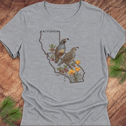 A heather gray short-sleeve t-shirt with a graphic print of two quail birds on a California state outline, surrounded by red berries, yellow flowers, and the word 'CALIFORNIA'. The shirt is displayed flat on a rustic wood background with pine branches in the corners.