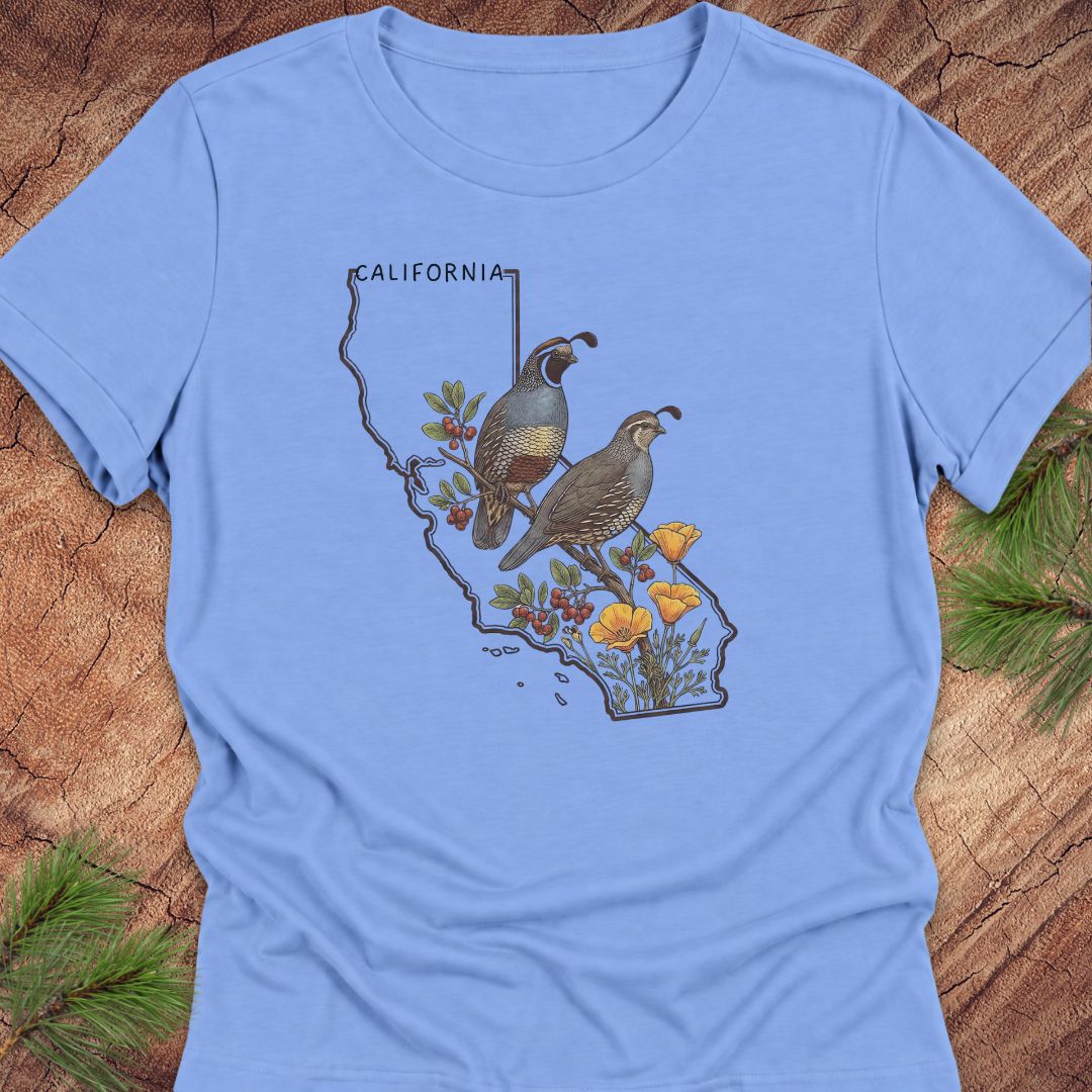 A light blue short-sleeve crewneck t-shirt laid flat on a rustic wood background with pine needles. The front of the shirt features a graphic print of the map of California decorated with two quail birds, red berries, and yellow wildflowers.