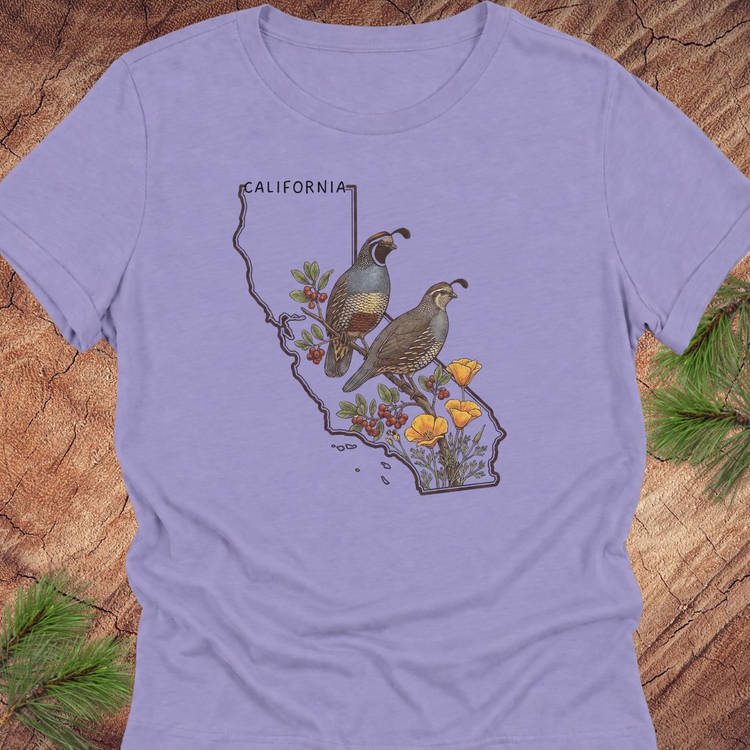 A lavender short-sleeve crewneck t-shirt featuring a graphic print of a California state outline filled with two quail birds, yellow flowers, and berries. The shirt is displayed flat on a rustic wood background with pine needles in the corners.