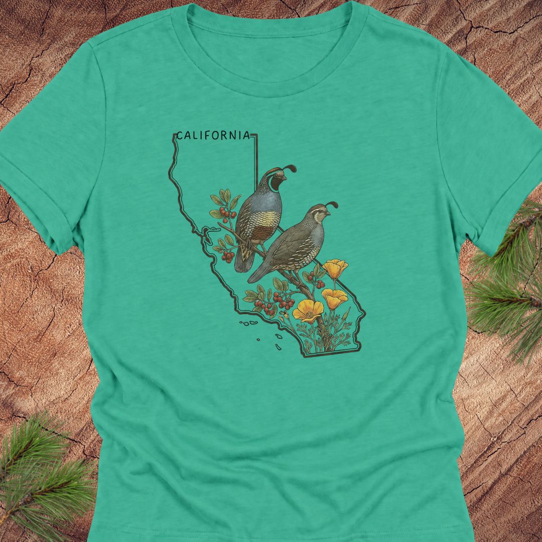 A teal short-sleeve crewneck t-shirt featuring a graphic print of a California map filled with two quail birds, yellow wildflowers, and a vertical 'CALIFORNIA' sign, laid flat on a rustic wood background with pine branches.