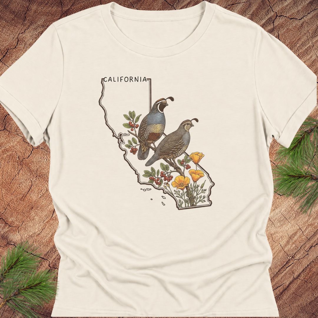An off-white short-sleeve t-shirt with a graphic print of two quail birds on a map of California filled with yellow flowers and berries, laid flat on a rustic wood background with pine needles.