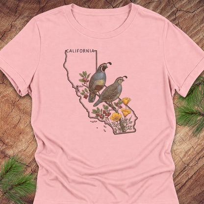 A light pink short-sleeve t-shirt with a graphic print of a California map outline featuring two quail birds, cactus plants, and yellow flowers. The shirt is laid flat on a rustic wood background with pine needles visible in the corners.