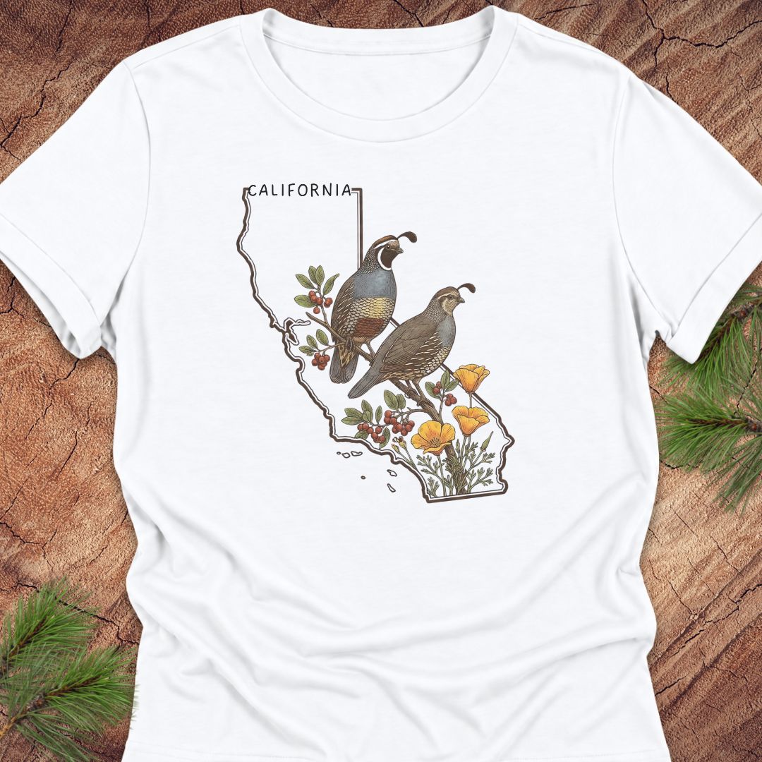 A white short-sleeve crewneck t-shirt laid flat on a rustic wood background with pine needles. The shirt features a graphic print of a map of California filled with two quail birds, green leaves, red berries, and yellow wildflowers.