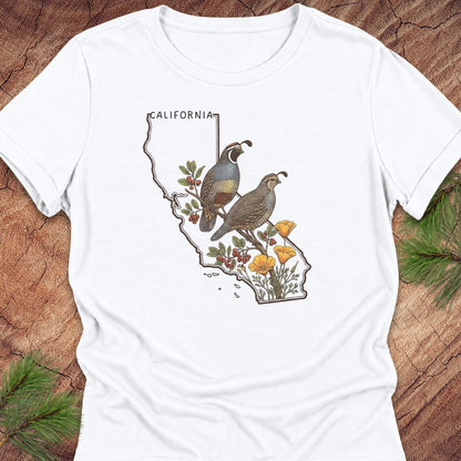 A white short-sleeve crewneck t-shirt laid flat on a rustic wood background with pine needles. The shirt features a graphic print of a map of California filled with two quail birds, green leaves, red berries, and yellow wildflowers.