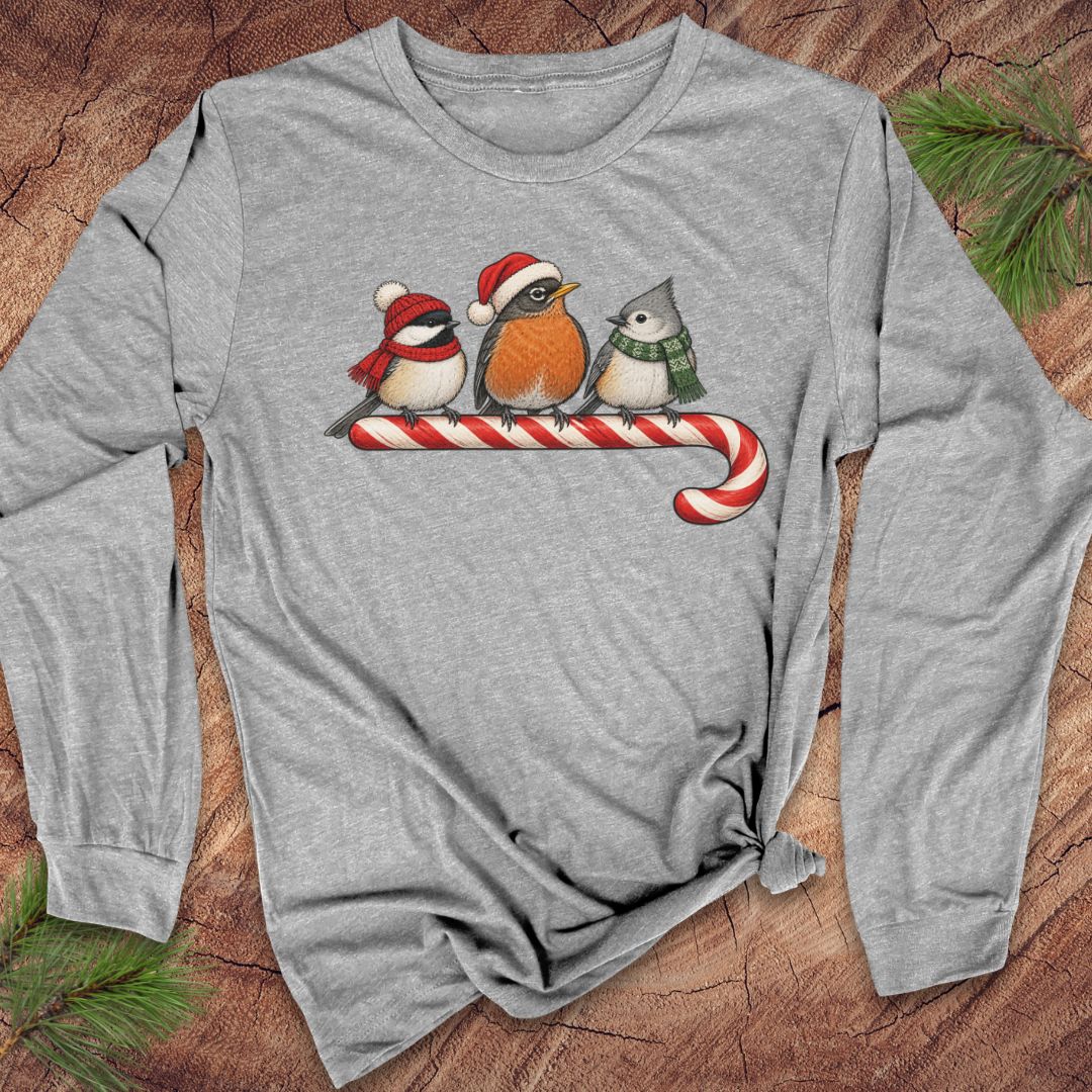 Athletic heather gray long-sleeve t-shirt with a Black-capped Chickadee, American Robin, and Tufted Titmouse in winter hats and scarves perched on a candy cane, laid flat on a rustic wood background with pine branches.