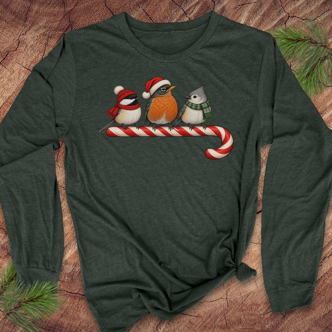Heather forest green long-sleeve t-shirt with a Black-capped Chickadee, American Robin, and Tufted Titmouse in winter hats and scarves perched on a candy cane, laid flat on a rustic wood background with pine branches.