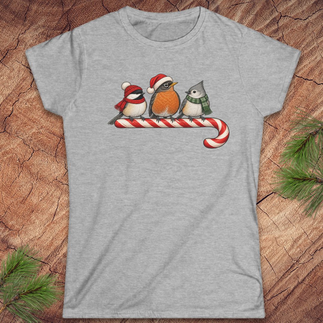 Sport grey women's t-shirt with a Black-capped Chickadee, American Robin, and Tufted Titmouse in winter hats and scarves perched on a candy cane, laid flat on a rustic wood background with pine branches.