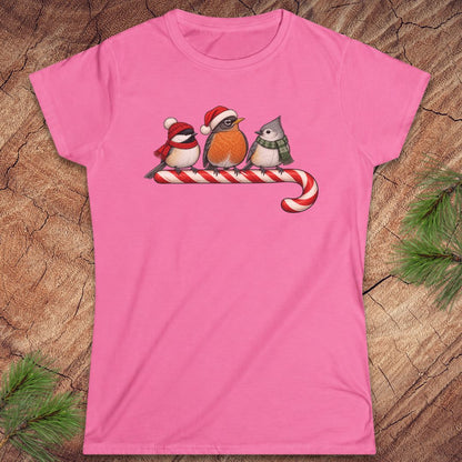 Azalea pink women's t-shirt with a Black-capped Chickadee, American Robin, and Tufted Titmouse in winter hats and scarves perched on a candy cane, laid flat on a rustic wood background with pine branches.