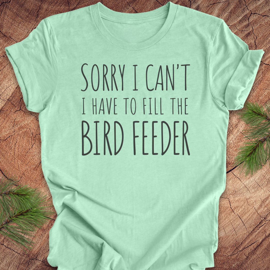 A mint green short-sleeve t-shirt with the text 'SORRY I CAN'T I HAVE TO FILL THE BIRD FEEDER' printed in black on the front, laid flat on a rustic wood background with pine branches in the corners.