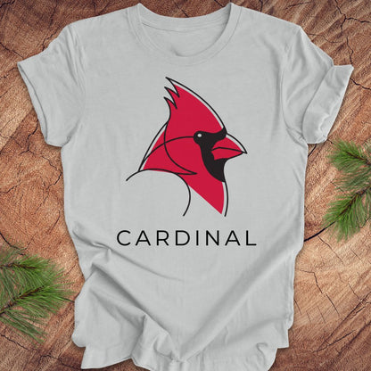 A light gray short-sleeve t-shirt laid flat on a rustic wood background, featuring a red cardinal bird graphic with black details and the word 'CARDINAL' printed below in black.