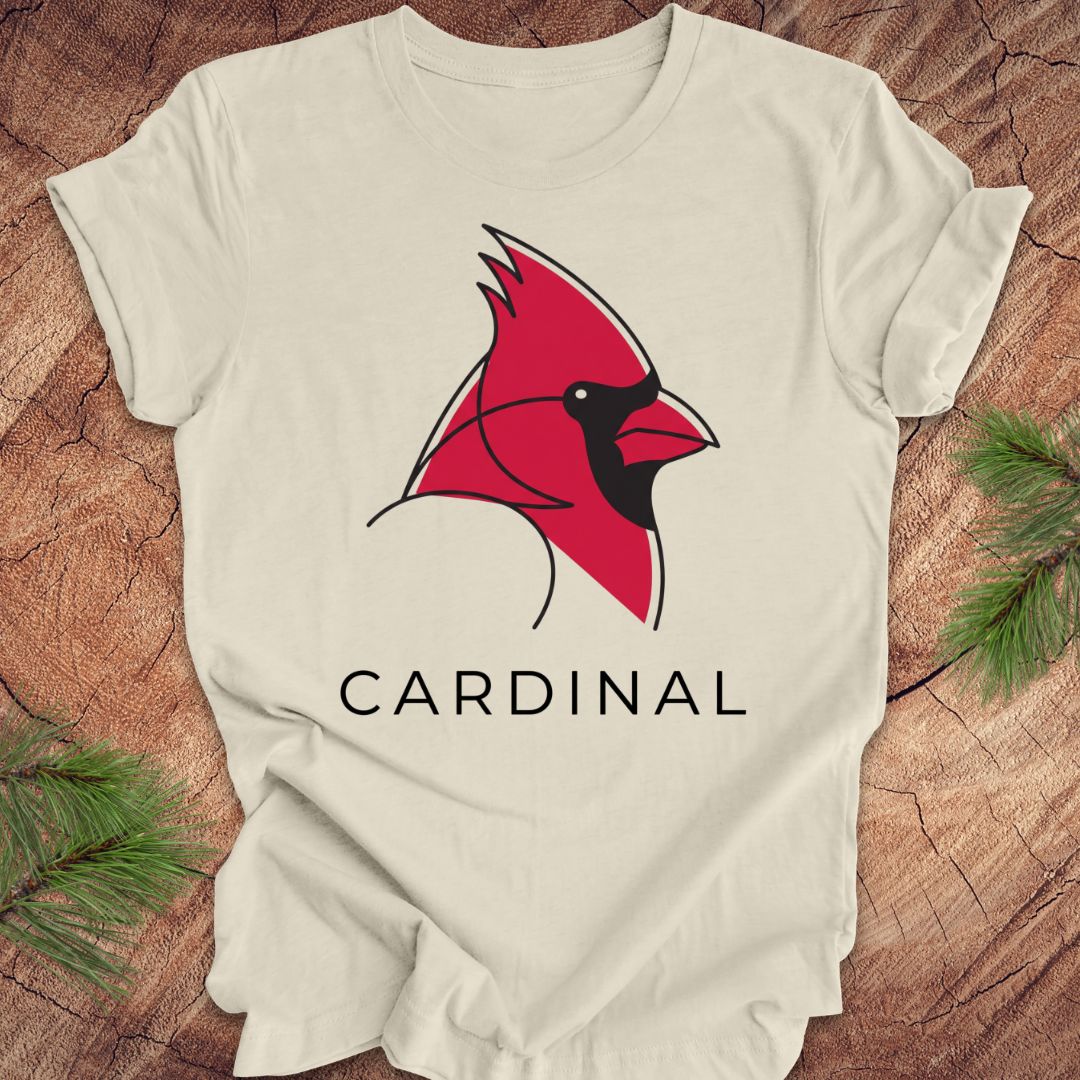 A beige short-sleeve t-shirt laid flat on a rustic wood background, featuring a stylized red cardinal bird graphic with a black beak and the word 'CARDINAL' printed below in black.
