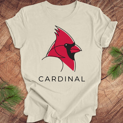 A beige short-sleeve t-shirt laid flat on a rustic wood background, featuring a stylized red cardinal bird graphic with a black beak and the word 'CARDINAL' printed below in black.