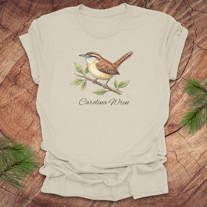 Beige t-shirt with a Carolina Wren illustration and text, 'carolina wren' on a wooden surface