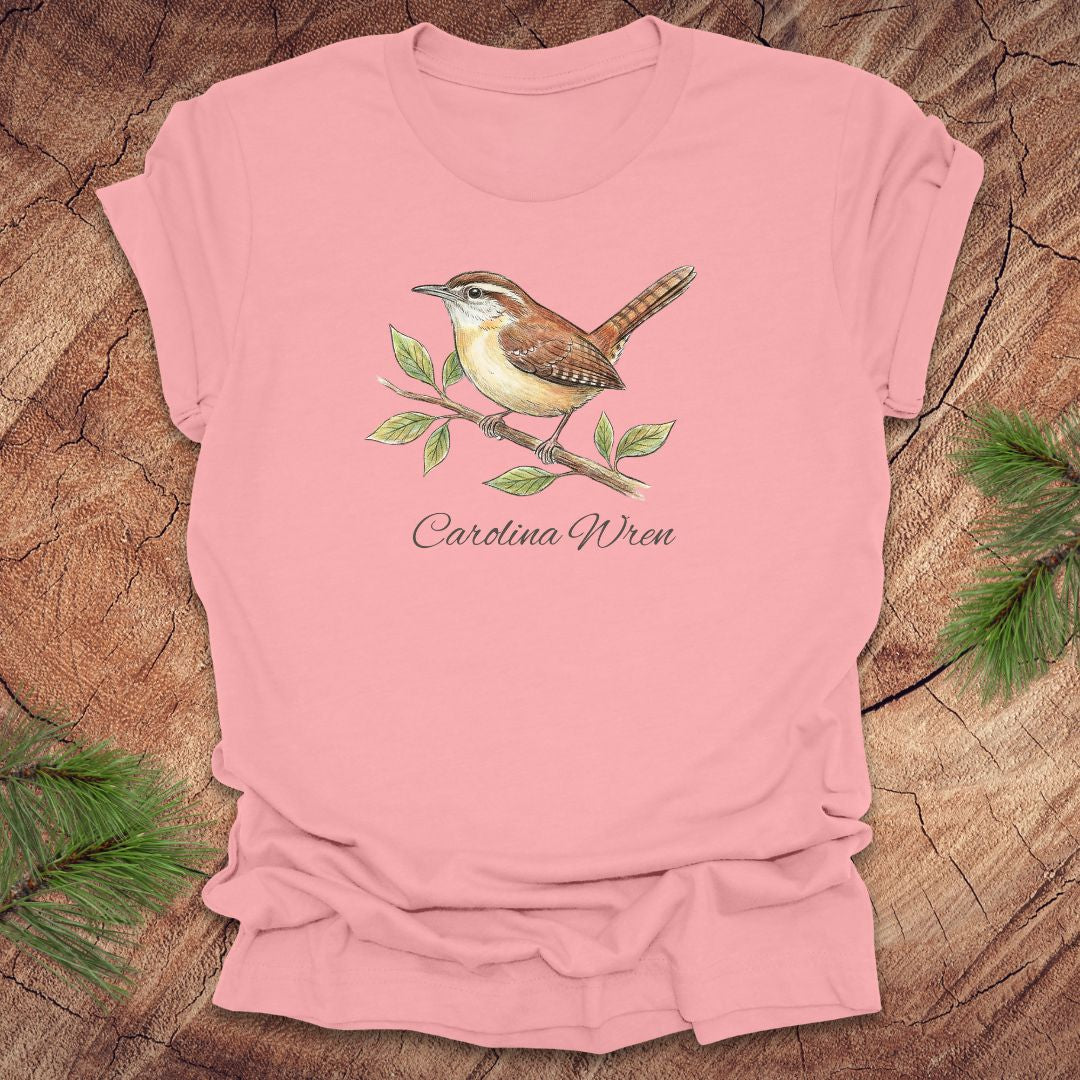 Pink t-shirt with a Carolina Wren illustration and text, 'carolina wren' on a wooden surface