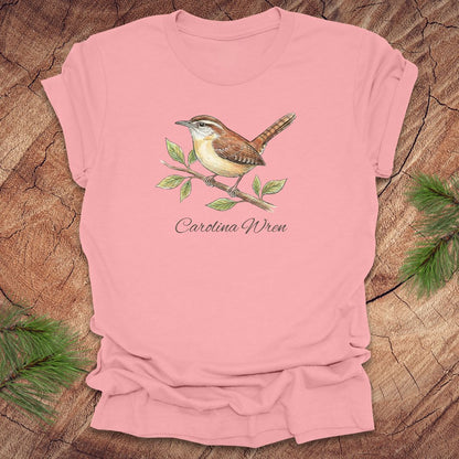 Pink t-shirt with a Carolina Wren illustration and text, 'carolina wren' on a wooden surface