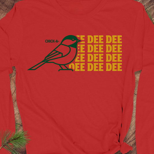 A red long-sleeve t-shirt with a graphic of a chickadee bird and the text 'CHICK-A-DEE DEE DEE' in yellow and green, displayed flat on a rustic wood background with a pine sprig in the corner.